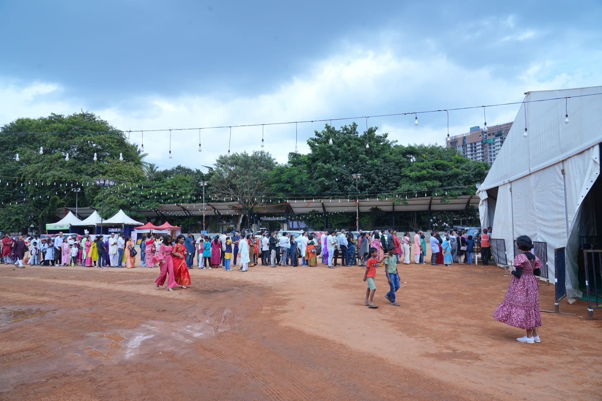 A long line of families in festive clothes waiting for bhog on the open Puja ground, with the tent on the right and strings of lights overhead