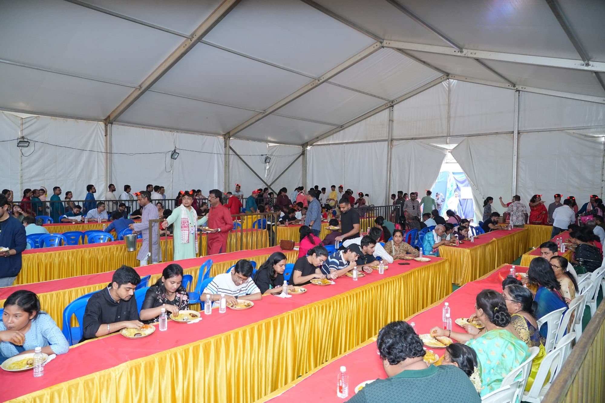 The dining hall at the 2025 Puja: long tables with red cloths and gold frills, families seated and eating bhog from leaf plates