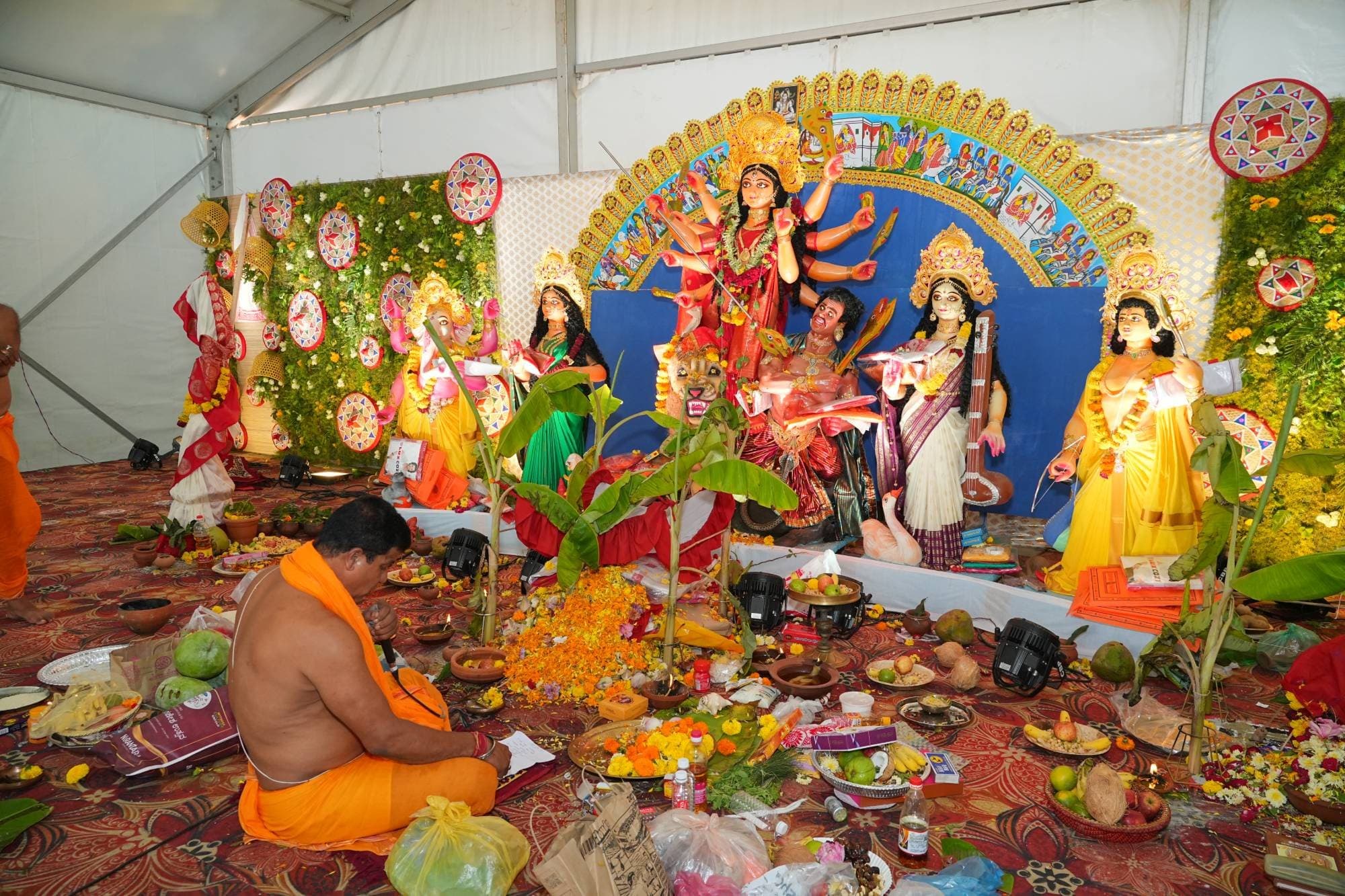 A priest in saffron sits reciting before Maa Durga and her family, with flowers, fruit, lamps and banana plants laid out in front of the idols
